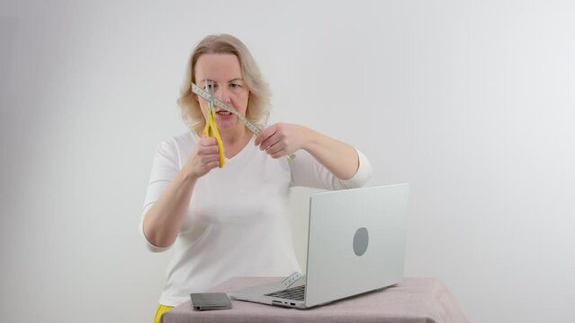 adult middle-aged woman is tired of diets she holds scissors in hands and cuts meter centimeter for Measure waist on table laptop in hands phone she angry opened mouth screams white background