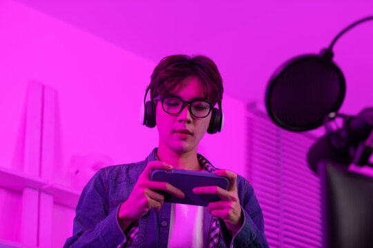 Professional Asian gamer playing and live streaming online game by smartphone in game room. Young man streaming video game on mobile phone with headphones and smiling, enjoying online gaming and tech.