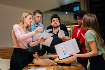 Portrait of calm male team leader sitting on office table in lotus position with closed eyes and meditating feeling calmness and serenity. Multiethnic annoyed fussy colleagues throw documents.