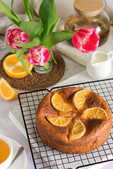 Cottage cheese orange cake decorated with orange slices on a wooden board and a stand, next to it is a mug of tea and a bouquet of flowers top view