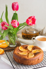 Cottage cheese-orange cake decorated with orange slices on a wooden board and stand, next to it is a mug of tea and a bouquet of flowers on the table