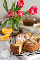 Cottage cheese-orange cake, decorated with orange slices and sprinkled with powdered sugar with a cut off piece, next to it is a mug of tea and a bouquet of flowers on the table