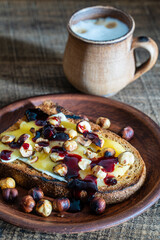 Bread toast with butter, roasted hazelnuts, honey, berry jam on plate and cappuccino coffee in a clay cup on wooden table, closeup
