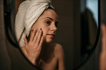 A Caucasian female wrapped in a towel standing in the bathroom after having a bath and applying...