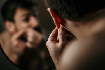 Obraz premium Shot of a young man examining his skin in the mirror at home. Health, skincare and man popping acne in face