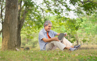 Naklejka na ściany i meble senior man with grey hair holding a book, smiling, sitting near a big tree in forest park. concept of elderly people lifestyle,hobbies,relaxing,health benefits in nature,wellbeing