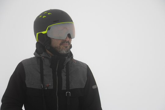 Hipster man is posing for a photo in akiing gear such as helmet and winter jacket. White snow background.