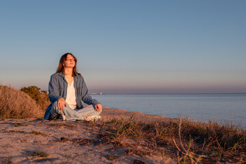 Young woman in jeans meditating on the seashore at sunset