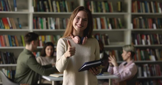 Positive fresh student girl holding notebook, standing in public college library with group of classmates studying in background, showing like hand sign at camera, making thumb up gesture