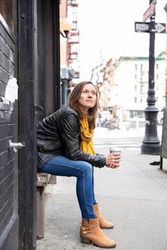 Side view of woman holding coffee while sitting on seat by building in city