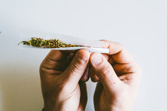 Cropped Hands Of Man Rolling Marijuana Joints In Paper Over White Table At Home