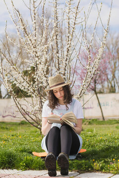 Full length of young woman reading book while sitting on skateboard at park
