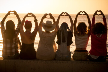 Group of women sitting against a golden sunset and doing heart symbol with hands and raised arms for friendship and love freedom concept. Women international day. Travel and vacation. Friends at beach