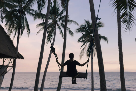 Rear view of young woman swinging on rope swing against coconut palm trees at beach during sunset