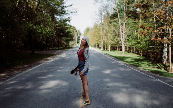 Full length portrait of cheerful woman with skateboard standing on road amidst trees
