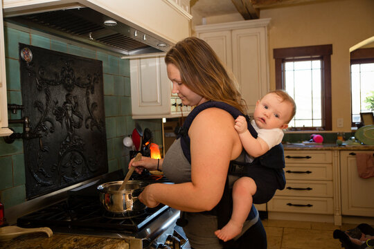 Side View Of Mother Carrying Son In Baby Carrier While Cooking Food At Kitchen