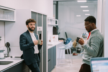 A young white male person talking to his black colleague while he is listening to him and looking at the cup