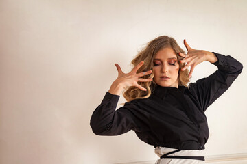 Lovely blonde lady in black shirt posing arms at white wall background, fan fingers, looking down. Studio shot of dancer woman with curly hair, dancing. Dance psychology concept. Copy text space ad