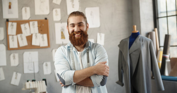 Close-up portrait shot of bearded hipster clothing designer working in his office, looking at camera and positively smiling - small business, fashion concept 