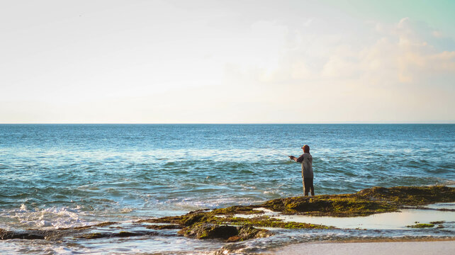 A man is fishing in the rocky sea in Bali, Indonesia. Angler fishing on a choppy beach during the day.
