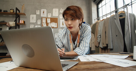 Hipster asian clothes designer working with her sketches, drawing them on graphics tablet,...