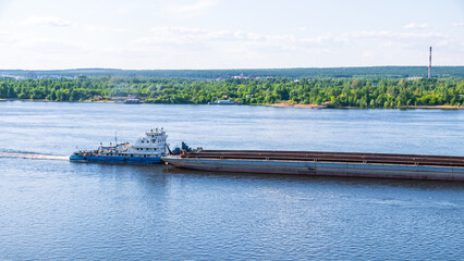 A tugboat pushes a large barge with cargo along a wide river. In summer, the tugboat goes down the...