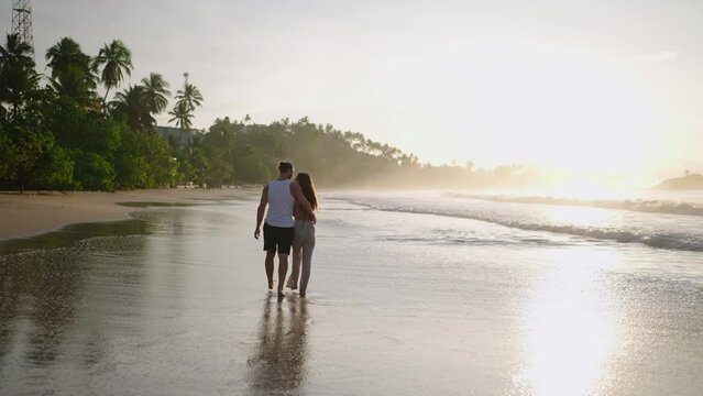 Silhouettes of a young happy couple holding hands and walking on the beach together enjoying summer back view. Boyfriend and girlfriend relax and walk at the seaside hugging and kissing at sunrise.
