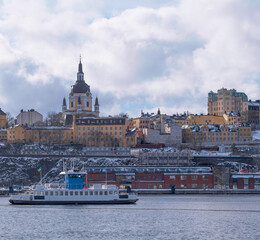 Fototapeta premium Harbor shuttle ferry passing the museum Fotografiska and the block at the church Katarina in the district Södermalm, a snowy day in Stockholm