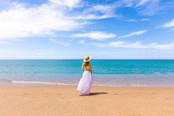 Cheerful woman in a straw hat standing alone on sandy beach, embracing the beauty and tranquility of tropical vacation. Clear ocean water with waves and sea breeze. Back view