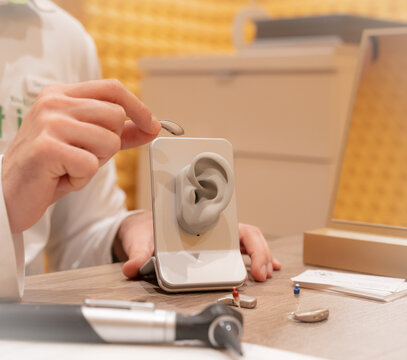 Doctor in his audiology office holding a hearing aid and in the other hand a model of a silicone ear. On his table is another hearing aid and medical devices.