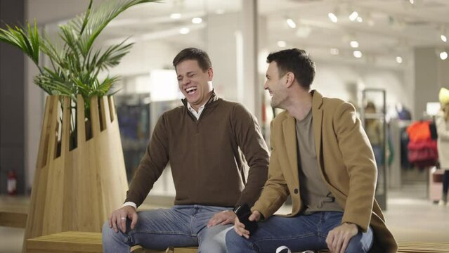 Two Young Friends Sitting On Chair In Shopping Mall, Talking And Smiling Loudly After Shopping. Shot In Slow-motion