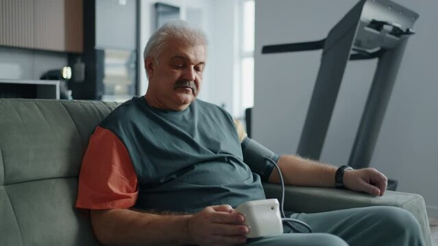 Aged Man Sitting On Couch At Home And Taking Blood Pressure By Electrical Sphygmomanometer