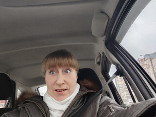 Portrait of adult cute funny blonde woman in warm clothes taking a selfie inside the car and driving it. The concept of a pleasant trip in spring, autumn, winter