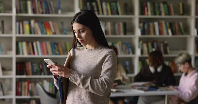 Serious young gen Z Latin student girl holding mobile phone, using app on smartphone in college library, typing, browsing, chatting, consulting internet, getting cheerful, happy, smiling