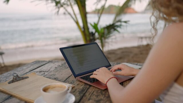 Female developer working on laptop by the ocean. Young woman freelancer coding at outdoor tropical cafe. Caucasian girl working remotely typing on computer at exotic location. Worldwide work concept.