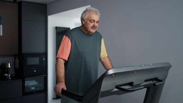 Medium Portrait Of Aged Man Walking On Treadmill And Listening To Music By In-Ear Headphones