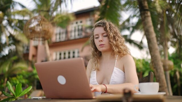 Young female developer working on laptop by the ocean. Woman freelancer coding at outdoor tropical cafe. Caucasian girl working remotely typing on computer at exotic location. Worldwide work concept.