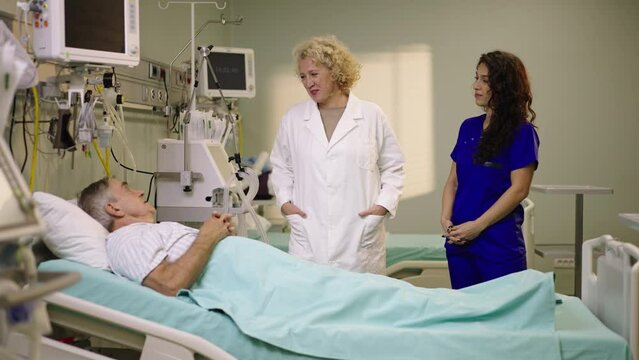 Professional mature doctor is on a round and talking to the patient who is lying on the bed in hospital. Shot in slow-motion