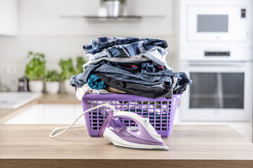 Big pile of wrinkled clothes pin a purple basket on a kitchen desk with an iron in front of it