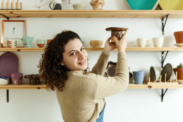 Happy young ceramist looking away thoughtfully while taking stock. Creative businesswoman managing a store with handmade ceramic products. Female entrepreneur running a successful small business