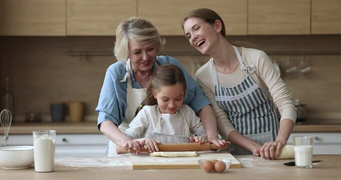 Happy mom and grandma teaching kid to bake, rolling dough for pastry together. Girl and women of three family generations cooking homemade bakery food, having fun, laughing at messy kitchen table