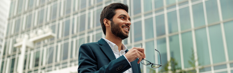 Smiling indian businessman in suit and glasses with laptop near office building