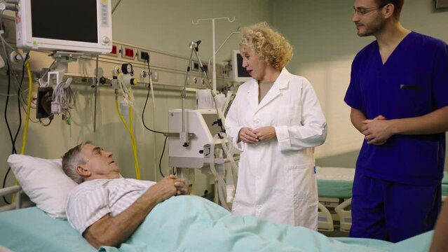 Mature female doctor in white apron explaining health condition of a patient in hospital ward. Shot in slow-motion