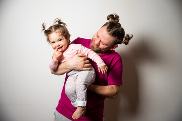 Young cute caucasian bearded father in bright pink t-shirt with two tails having fun with his little smiling daughter with two tails and finger into mouth
