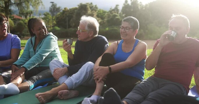 Multiracial senior people having fun drinking hot tea after workout exercises outdoor with city park in background - Healthy lifestyle and joyful elderly lifestyle concept