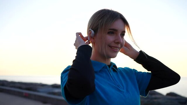 positive cute sporty smiling female puts on bone conduction headphones in sunset