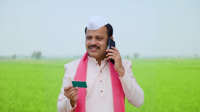 Village indian farmer talking on mobile phone by holding credit or green card at paddy farmland - concept of secure payment, banking or financial and modern farming.