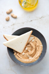 Dark-grey bowl with peanut butter and white bread slices on a light-beige marble background, vertical shot, high angle view