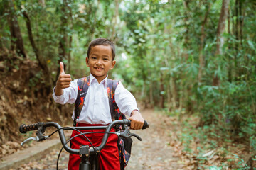 Fototapeta na wymiar a male elementary student in uniform riding his bike through the country road with thumb up and a lot of tree around him