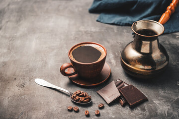 Black coffee drink in a clay cup, turkish jezve coffee pot, chocolate pieces and coffee beans in a spoon on dark background
